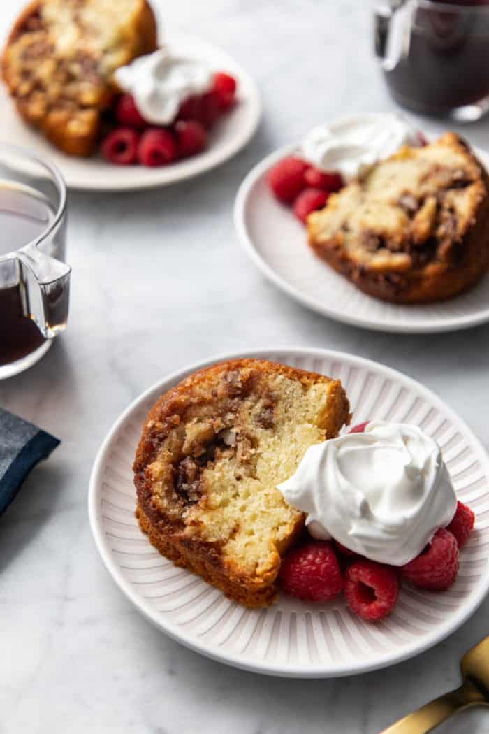 Easy coffee cake, fresh raspberries, and whipped cream on a white plate. Two more plates with the same thing can be seen in the background.