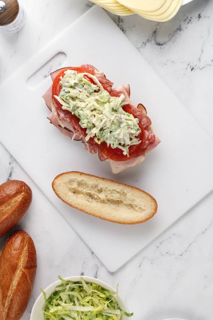 Salad, tomato, and deli meats layered onto a grinder sandwich roll on a white cutting board.