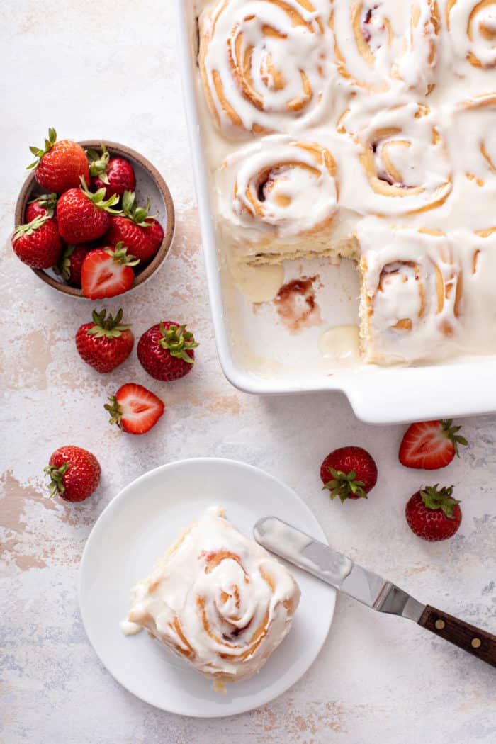 Offset spatula lifting a sweet roll onto a white plate from the baking dish of sweet rolls.