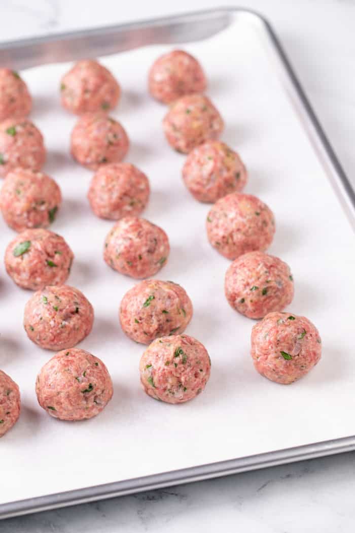 Portioned and rolled meatballs on a lined baking sheet, ready to be baked.