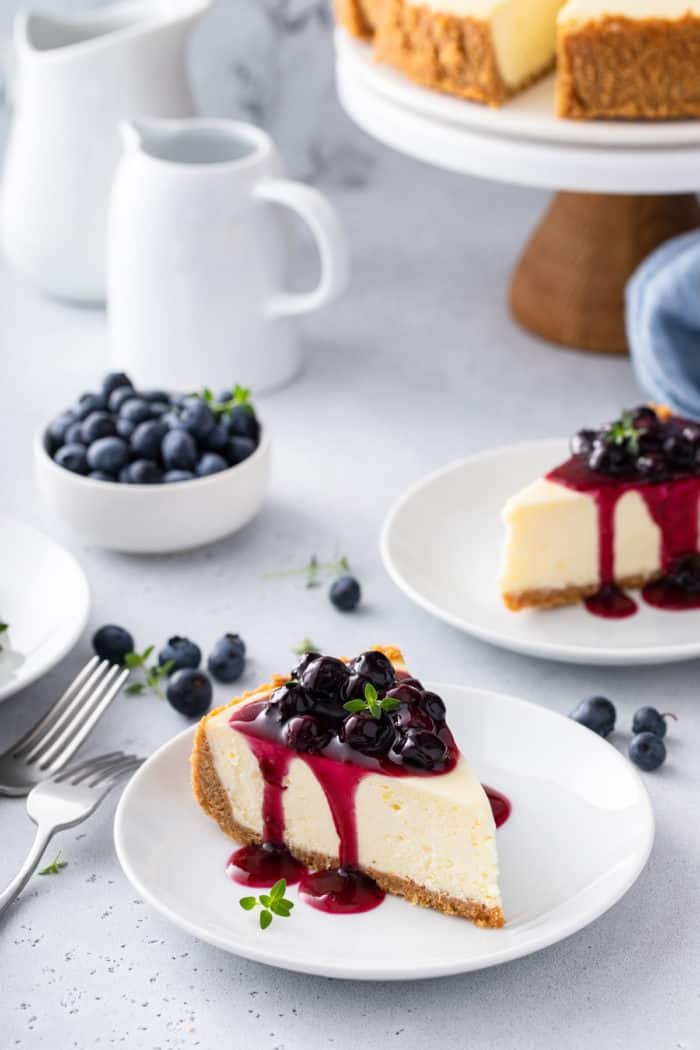 Two white plates with slices of blueberry cheesecake. A cake plate with the rest of the cheesecake can be seen in the background.