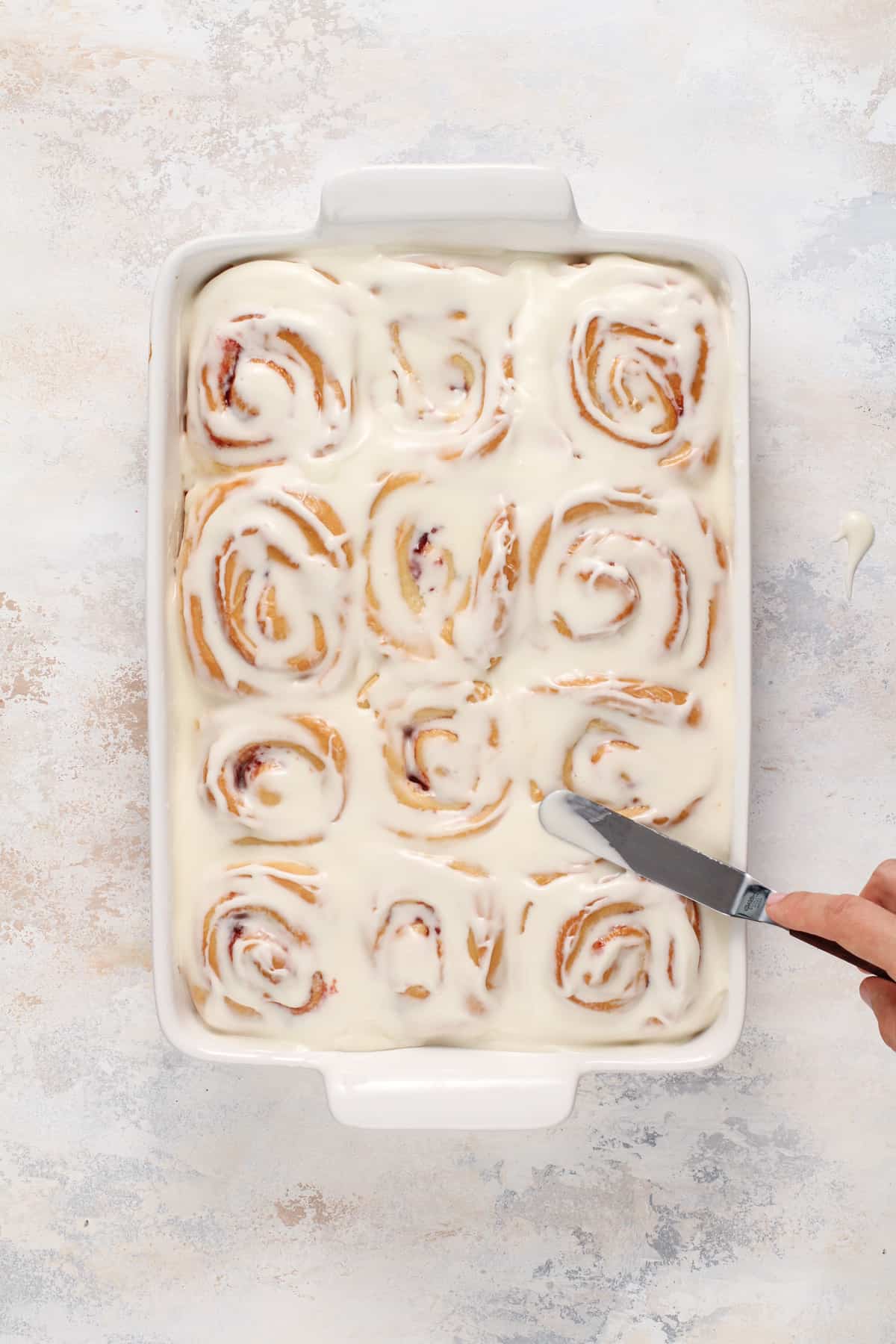 Hand spreading cream cheese frosting onto strawberry rolls with an offset spatula.