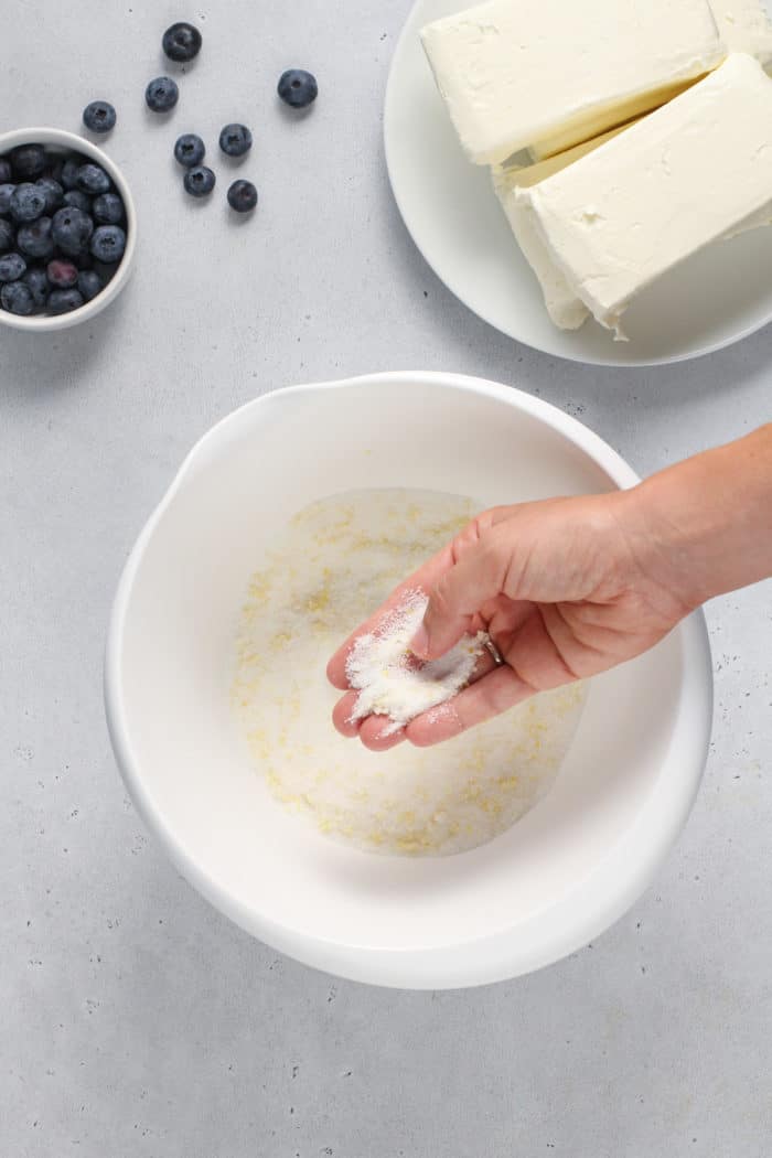 Sugar and lemon zest being mixed together with a hand in a white bowl.