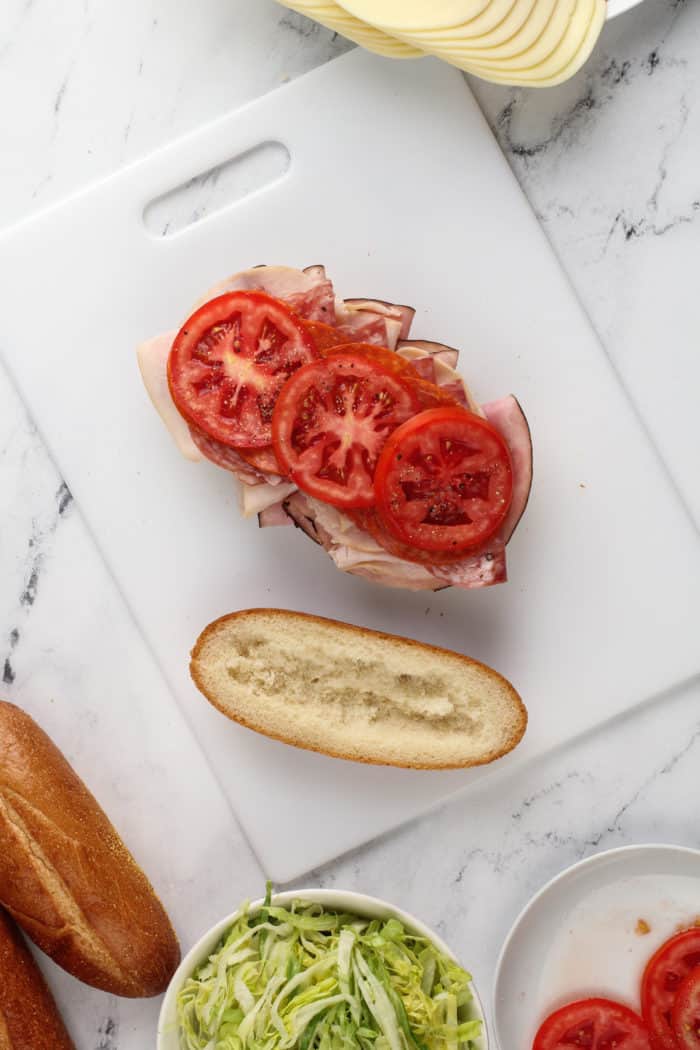 Sliced tomatoes on top of deli meats layered onto a grinder sandwich roll.