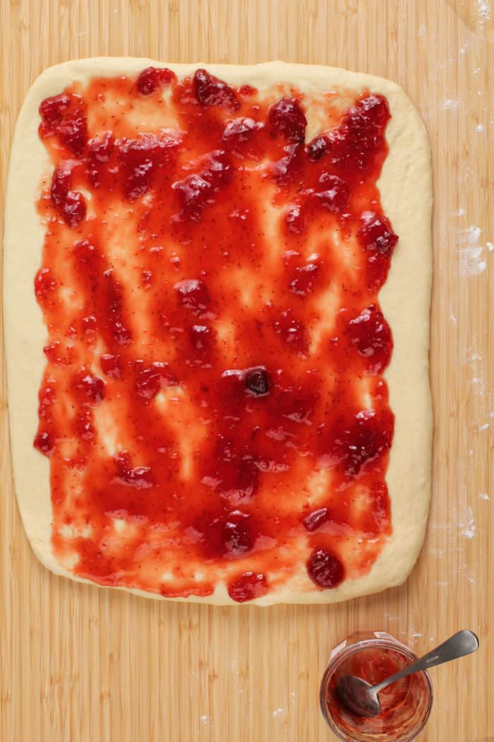 Strawberry jam spread over rolled-out cinnamon roll dough on a wooden board.