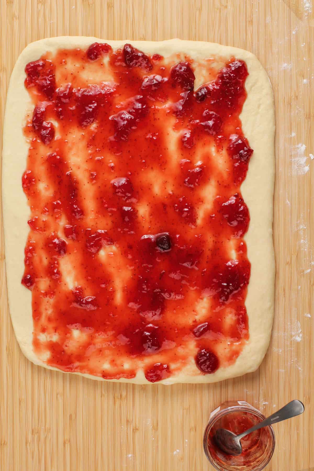 Strawberry jam spread over rolled-out cinnamon roll dough on a wooden board.