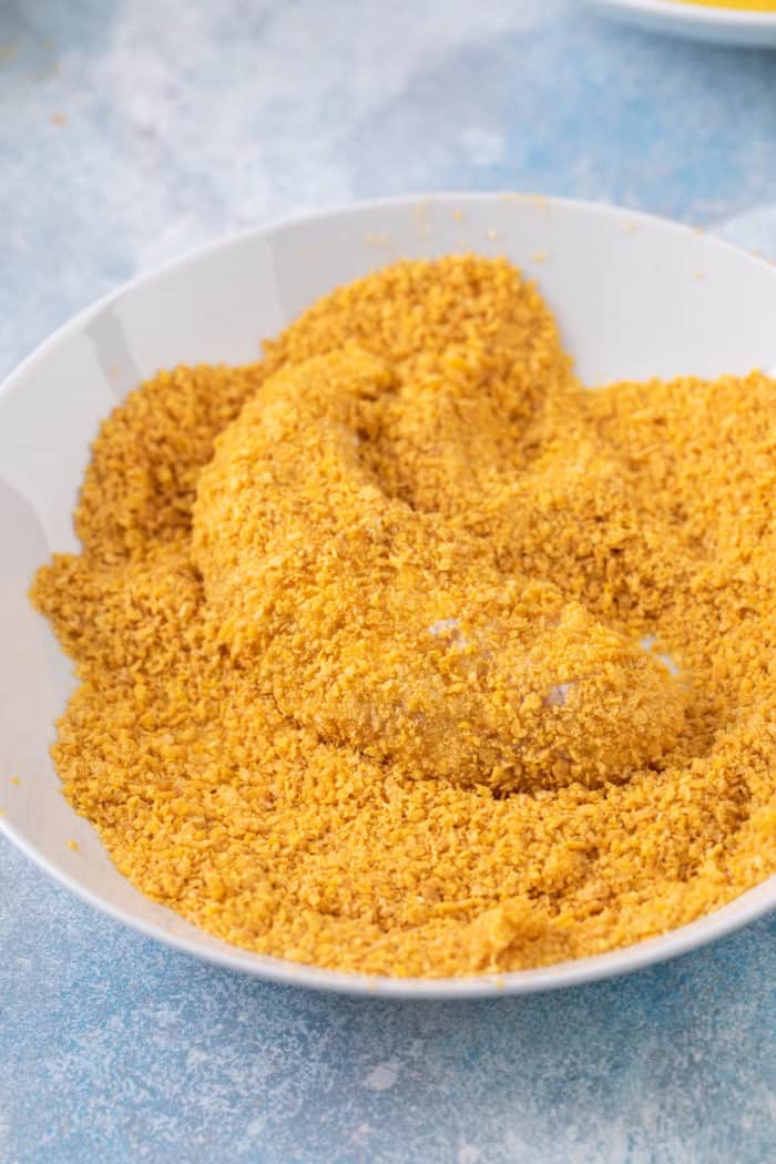 Chicken tender being dredged in corn flake crumbs in a shallow bowl.