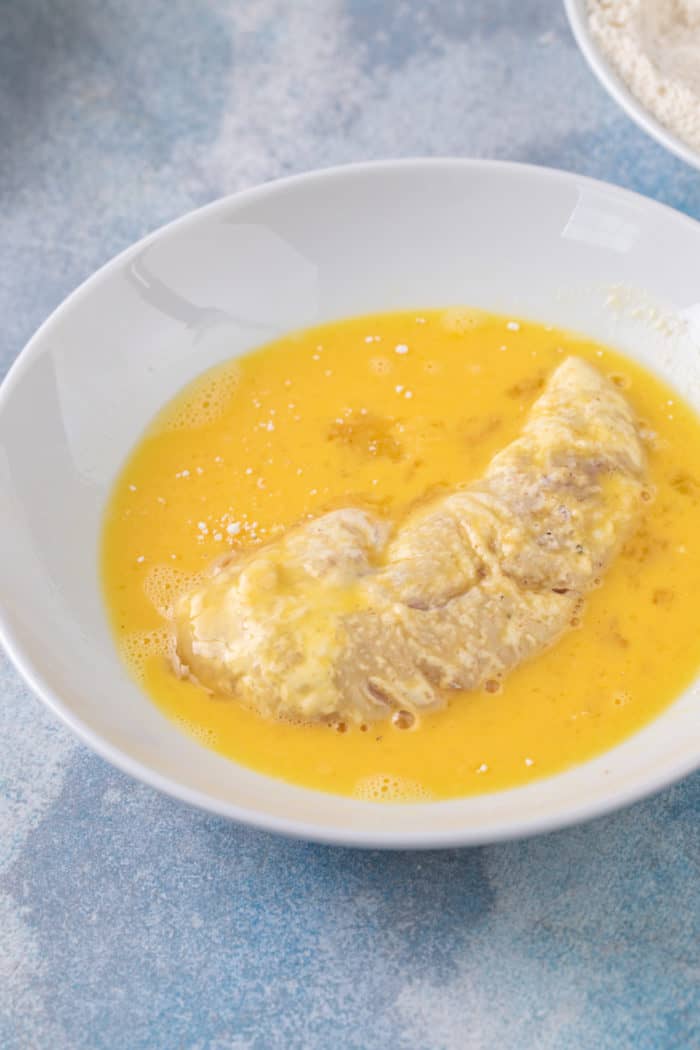 Chicken tender being dipped into egg wash in a shallow bowl.