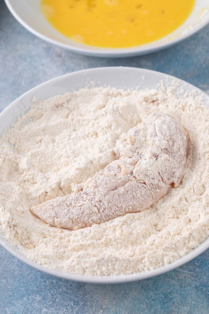 Chicken tender being dredged in seasoned flour in a shallow bowl.