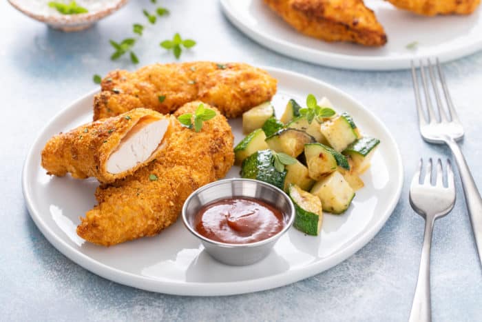 White plate containing air fryer chicken tenders, cooked zucchini, and a ramekin of barbecue sauce.