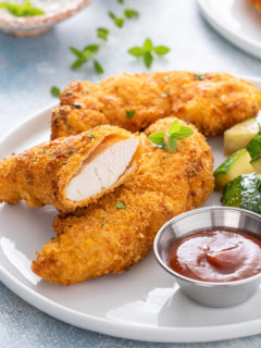 Three air fryer chicken tenders on a white plate next to a ramekin of barbecue sauce. One of the tenders is cut in half.