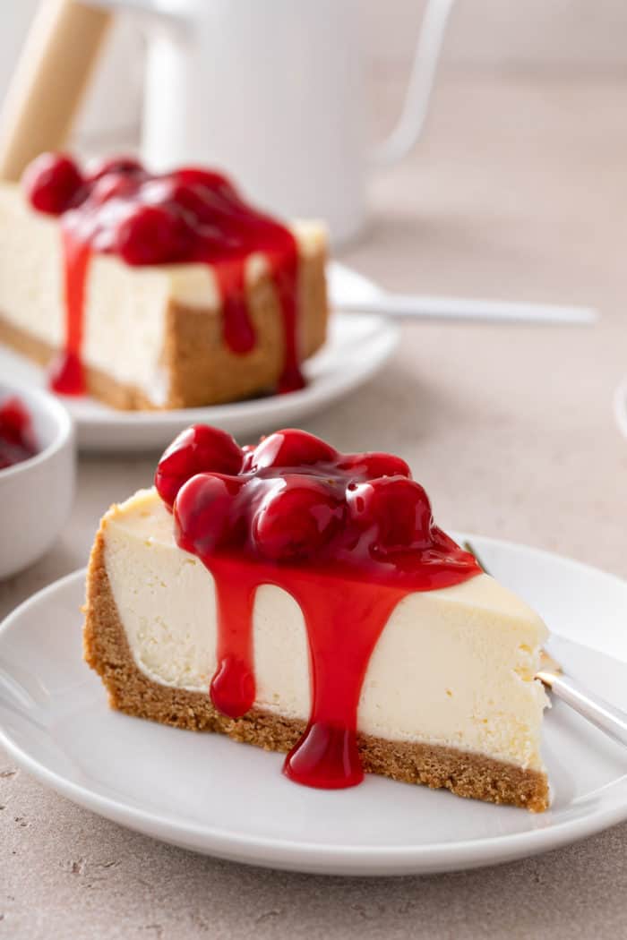 Close up view of the side of a slice of cherry cheesecake, set on a white plate. A second plate of cheesecake is in the background.