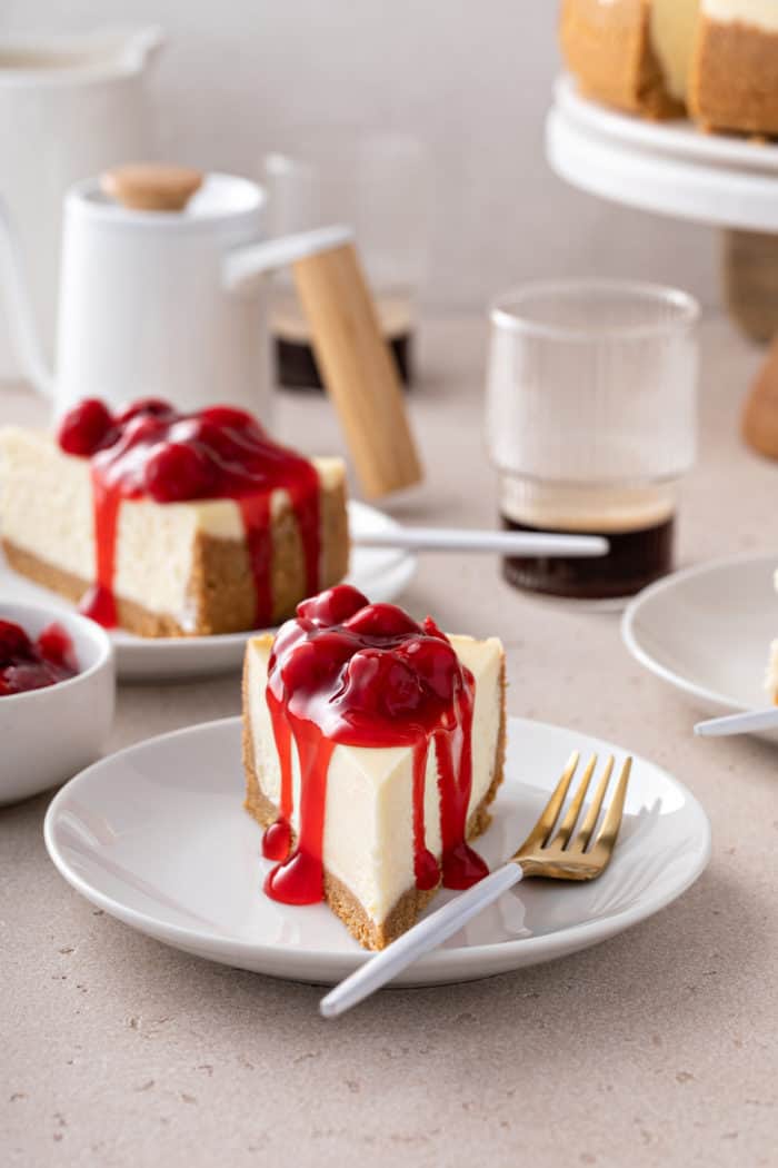 Slice of cherry cheesecake on a white plate, facing the camera. A second slice is visible in the background.