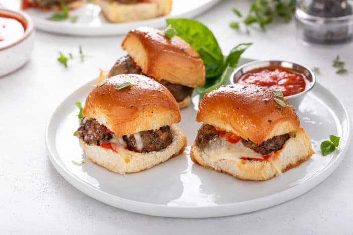 Three meatball sliders and a bowl of marinara sauce on a white plate.