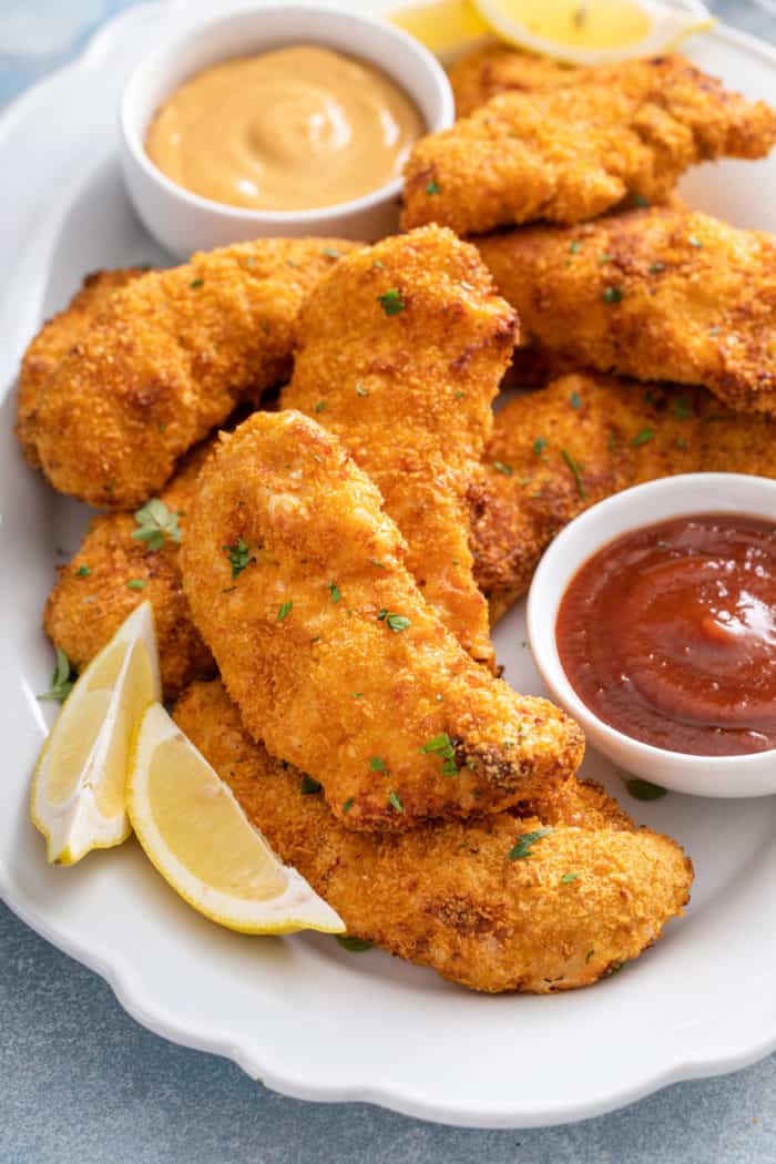 Close up of air fryer chicken tenders on a platter next to honey mustard sauce, barbecue sauce, and slices of lemon.