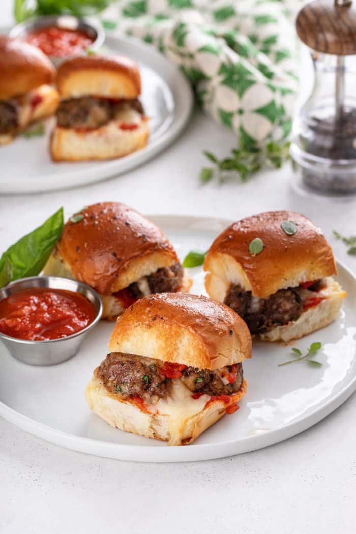 Three meatball sliders arranged on a white plate next to a small ramekin of marinara sauce.