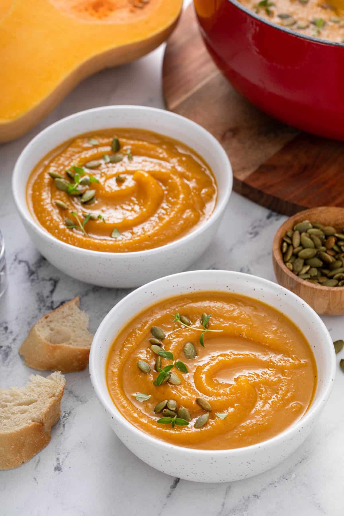 two bowls of butternut squash soup next to a pot of the soup and a bowl of pumpkin seeds