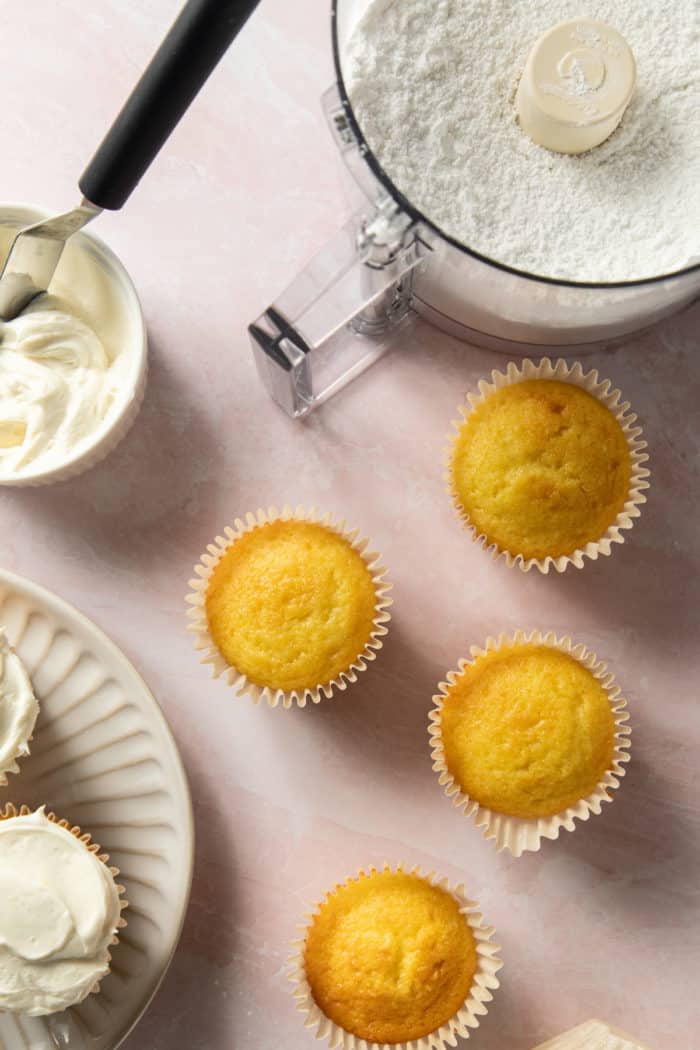 Bowl of a food processor with powdered sugar on a countertop next to cupcakes and a small bowl of vanilla frosting.