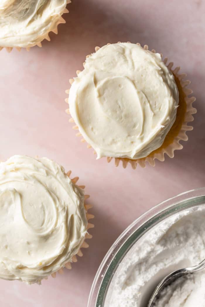 Frosted vanilla cupcakes set on a pink countertop.