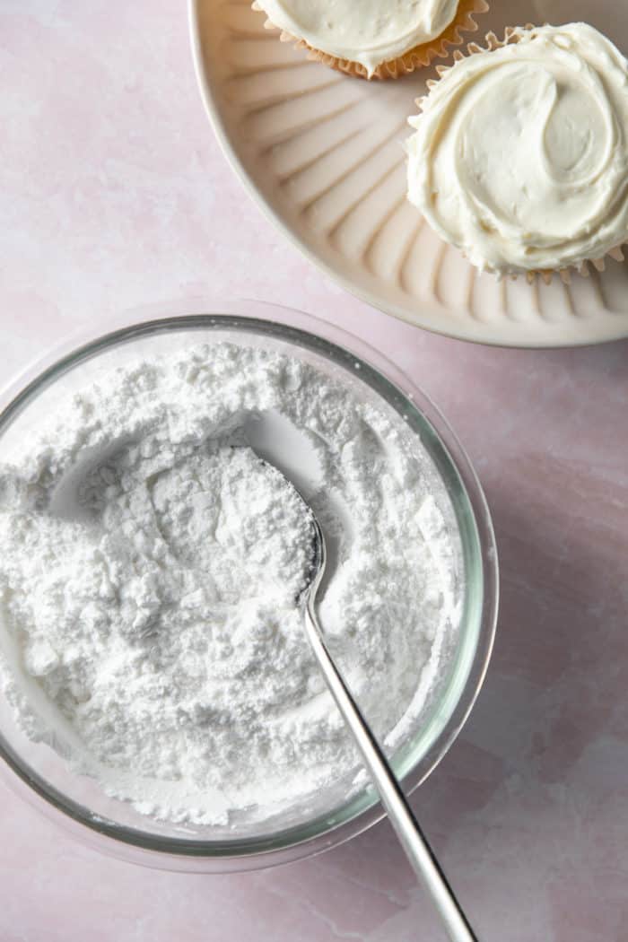 Bowl of powdered sugar set next to a plate of frosted cupcakes.