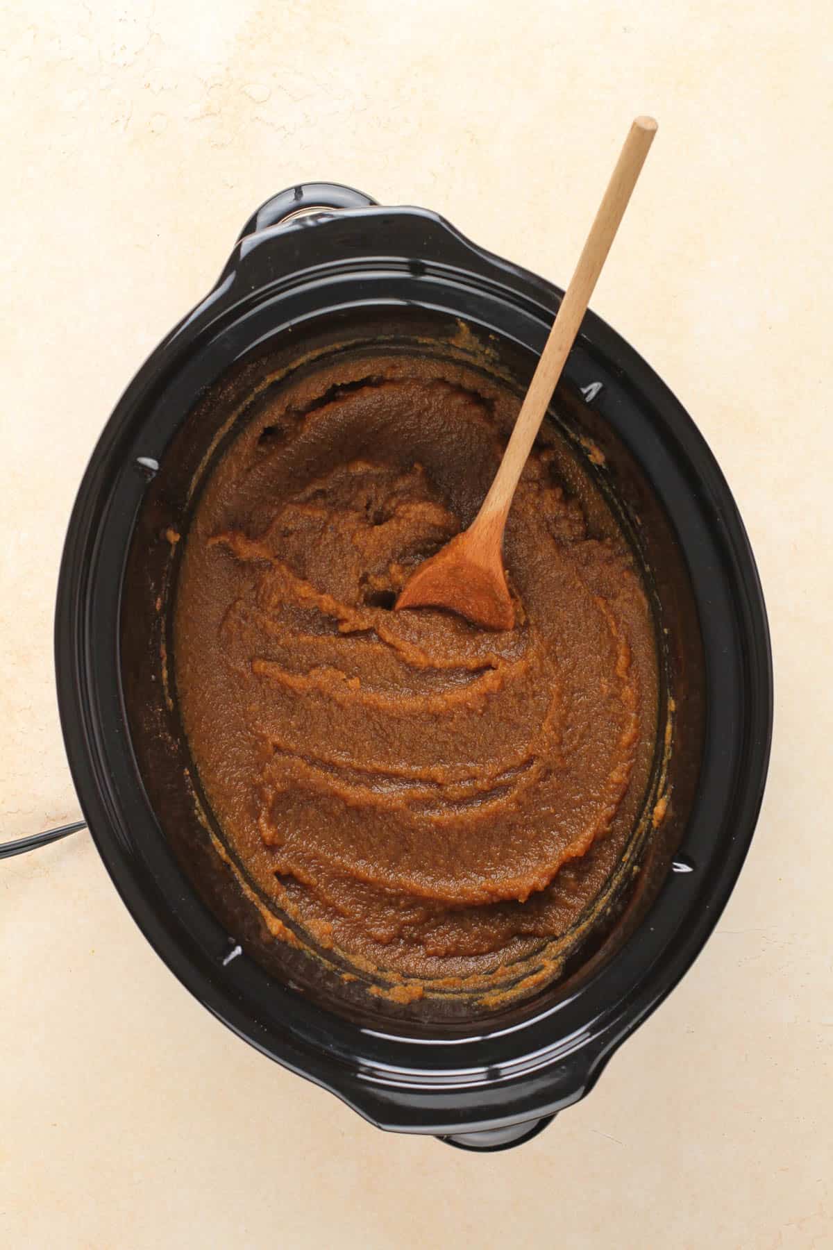a slow cooker containing cooked pumpkin butter and a wooden spoon