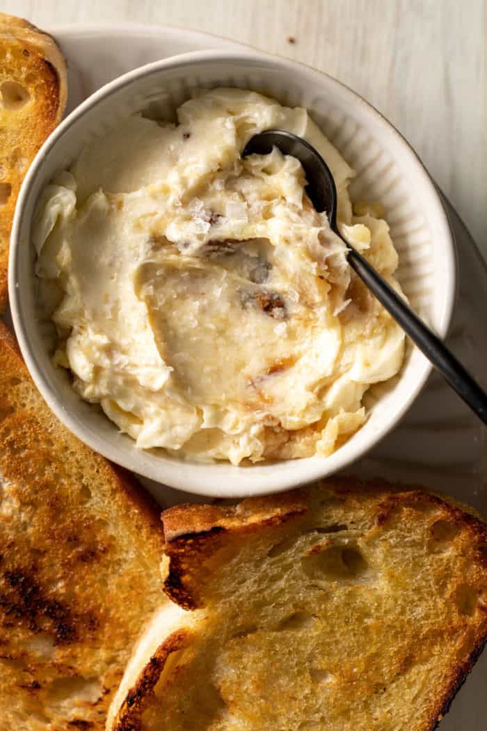 Bowl of roasted garlic butter next to slices of toasted baguette.