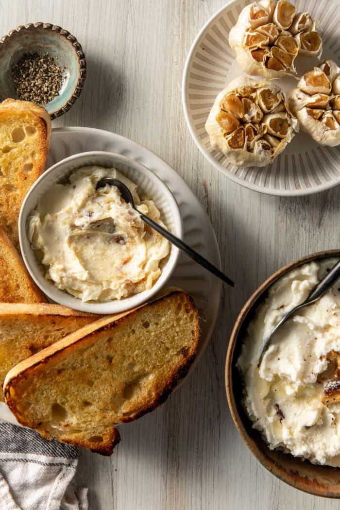 Roasted heads of garlic on a plate next to plates of garlic mashed potatoes and roasted garlic butter and toasted baguette.