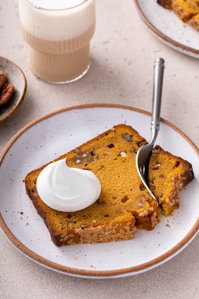 Fork cutting into a slice of brown butter pumpkin bread, topped with whipped cream, on a cream plate.