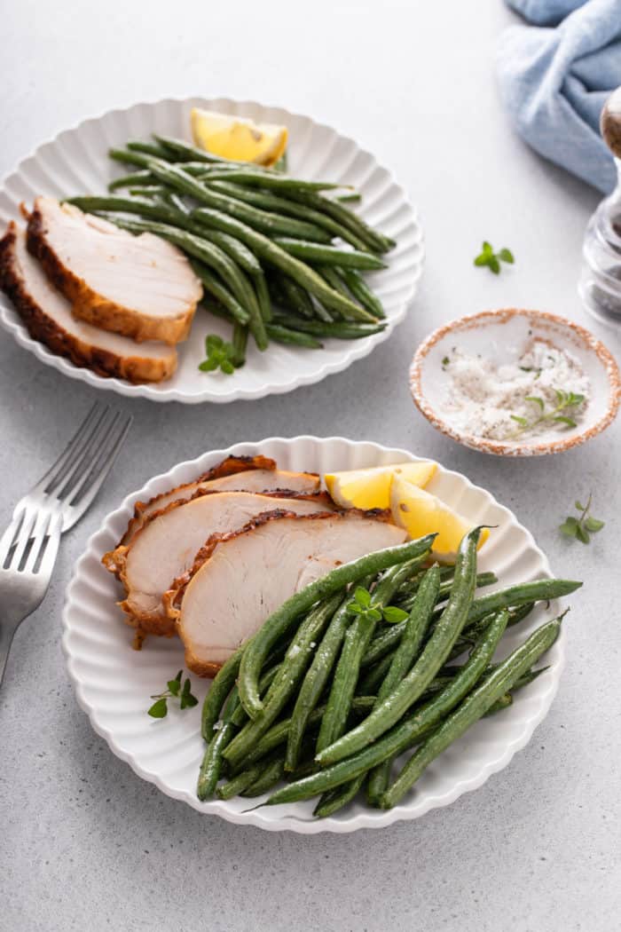 Air fryer green beans next to slices of roasted chicken and wedges of lemon on a white plate.