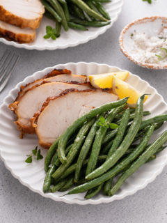 Air fryer green beans on a white plate next to roasted chicken and slices of lemon.