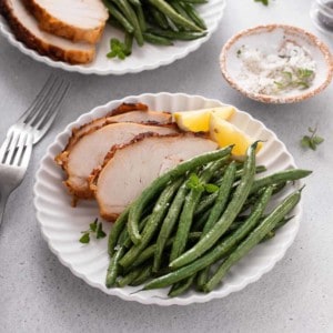 Air fryer green beans on a white plate next to roasted chicken and slices of lemon.