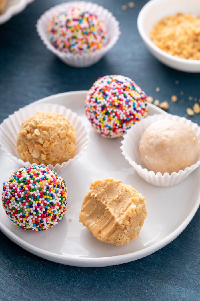 Five peanut butter balls rolled in a variety of toppings on a white plate. One ball has a bite taken out of it.
