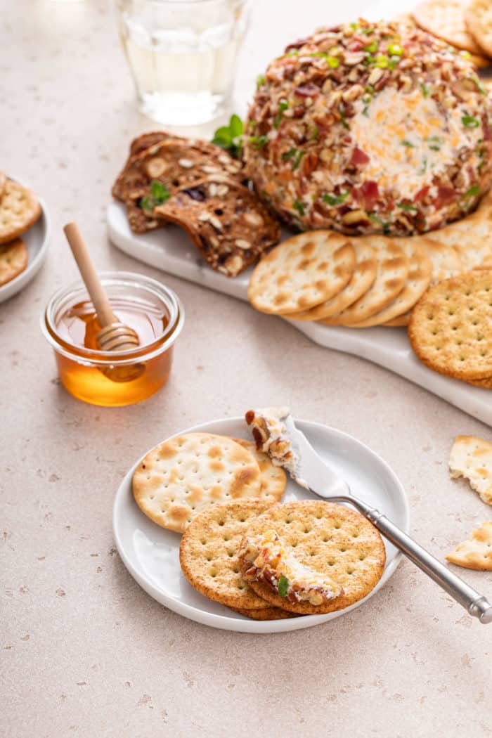 Small white plate with three crackers and a bit of bacon ranch cheese ball on it. A platter with the rest of the cheeseball is in the background.