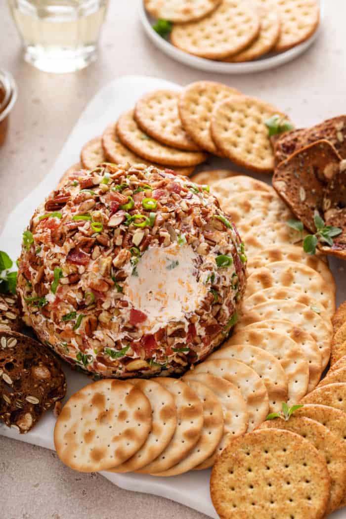 Bacon ranch cheese ball surrounded by crackers on a white platter. A bite has been taken from the side of the cheese ball.