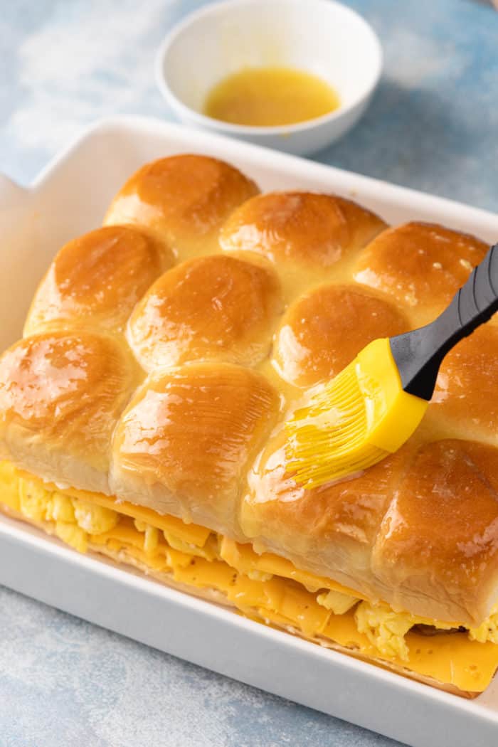 Melted butter being brushed over the top of unbaked breakfast sliders in a white baking dish.