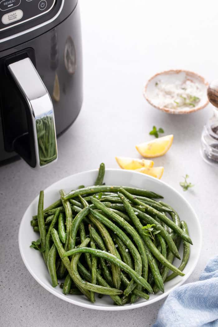 Bowl of air fryer green beans topped with flaky sea salt next to an air fryer.