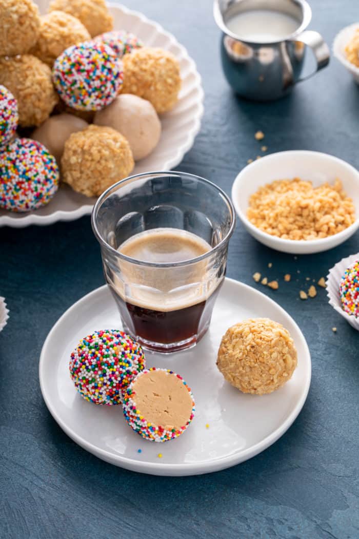 A cup of espresso set on a white plate next to two peanut butter balls. One ball is cut in half.