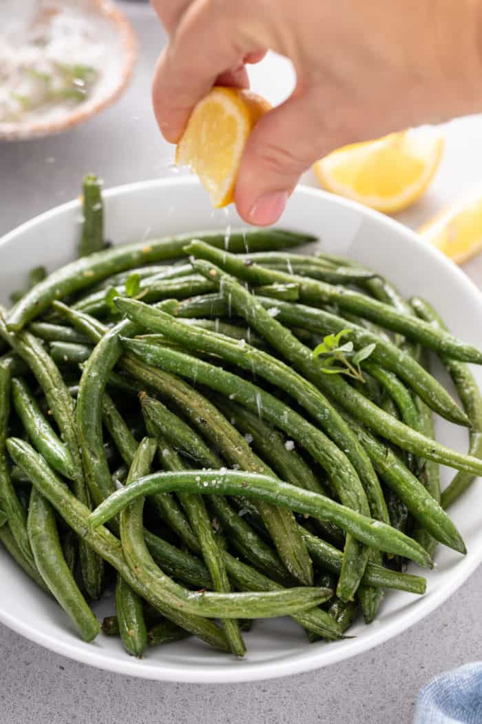 Lemon being squeezed over air fryer green beans in a white serving bowl.