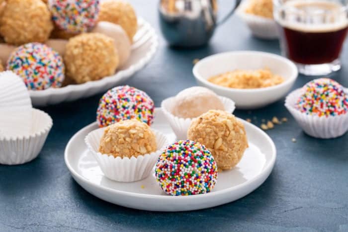 Five peanut butter balls on a white plate. Toppings on the peanut butter balls include rainbow nonpareil sprinkles, chopped nuts, and powdered sugar.
