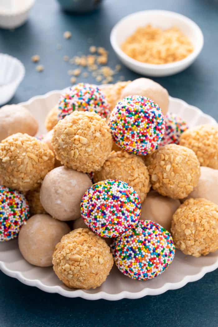 White platter filled with peanut butter balls rolled in chopped nuts, rainbow nonpareil sprinkles, and powdered sugar.