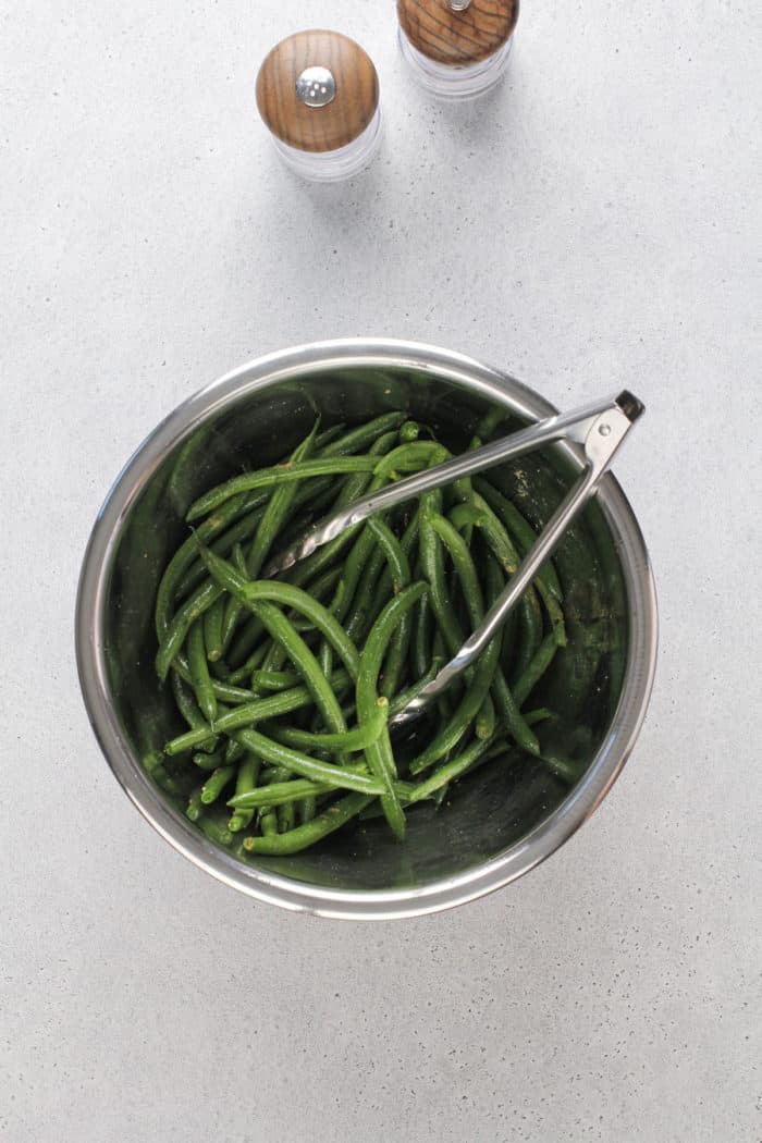 Tongs tossing fresh green beans with oil and seasoning in a metal bowl.
