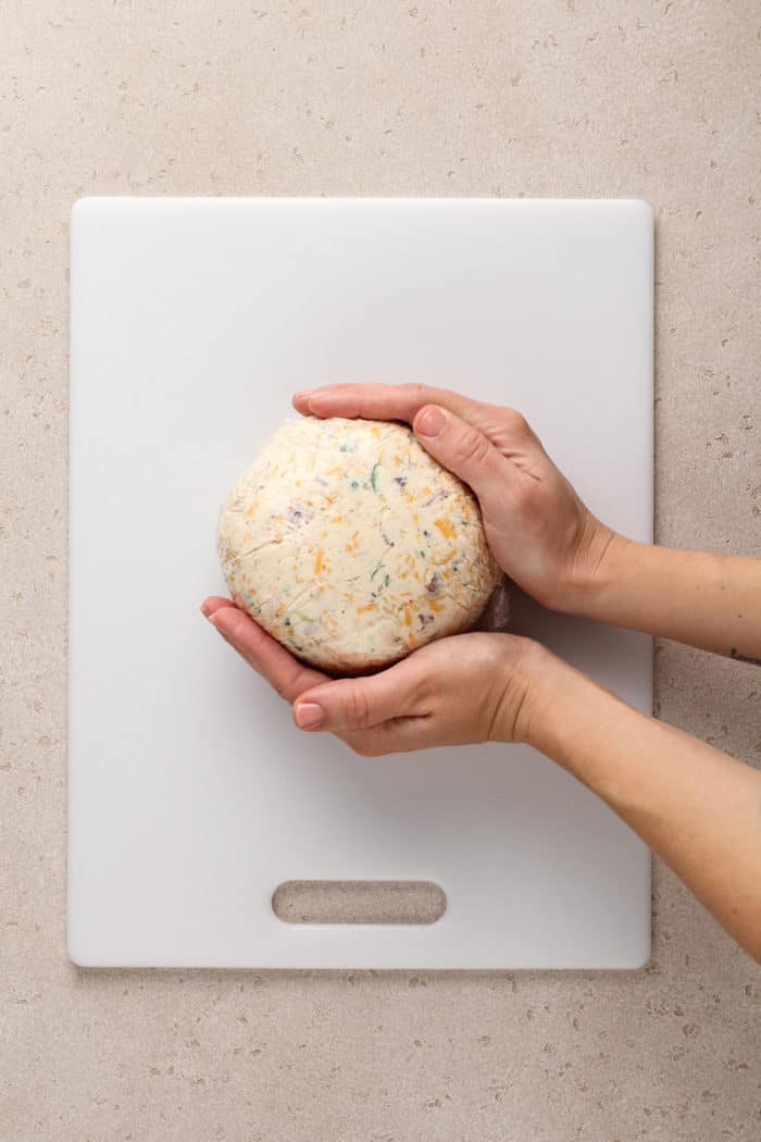 Hands shaping bacon ranch cheese ball into a ball on a white cutting board.
