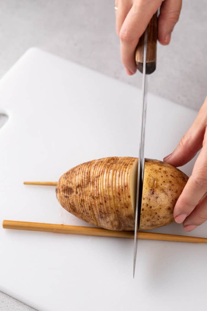 Knife cutting slices into a potato that is stabilized between two wooden chopsticks on a white cutting board.