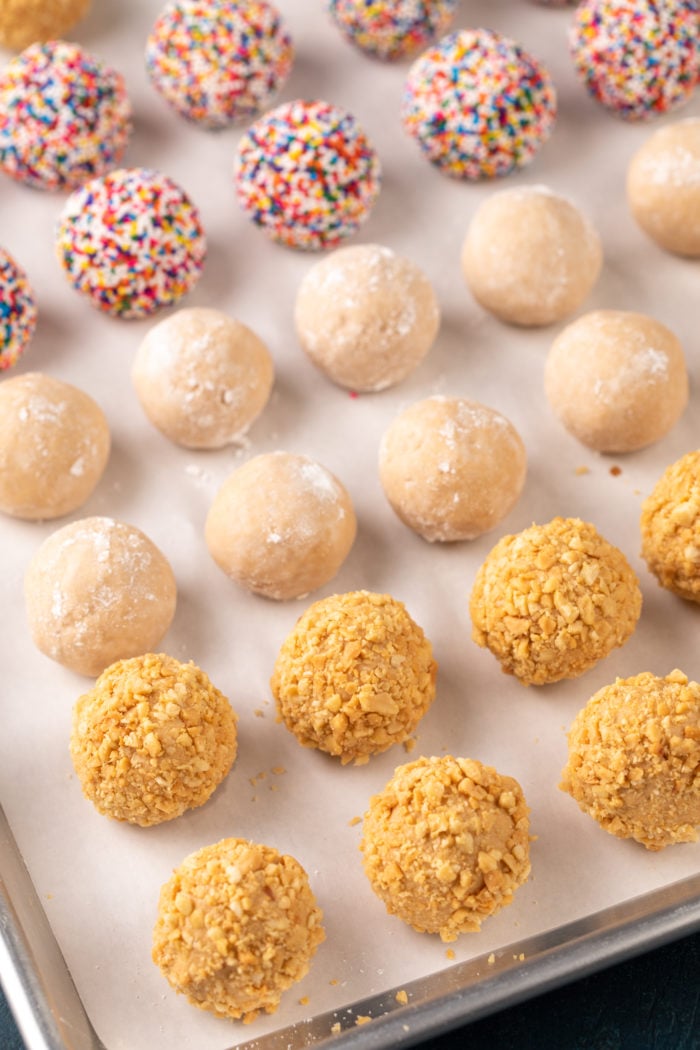 Parchment-lined sheet pan with peanut butter balls rolled in nuts, powdered sugar, and rainbow nonpareil sprinkles.