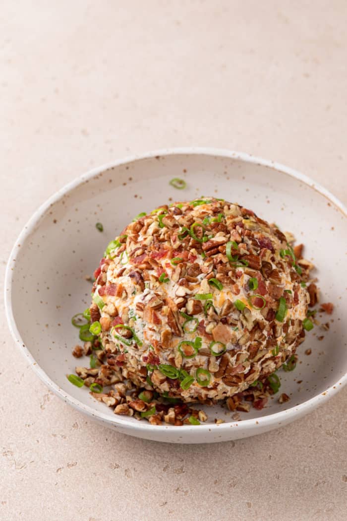 Bacon ranch cheese ball being rolled in toppings in a bowl.