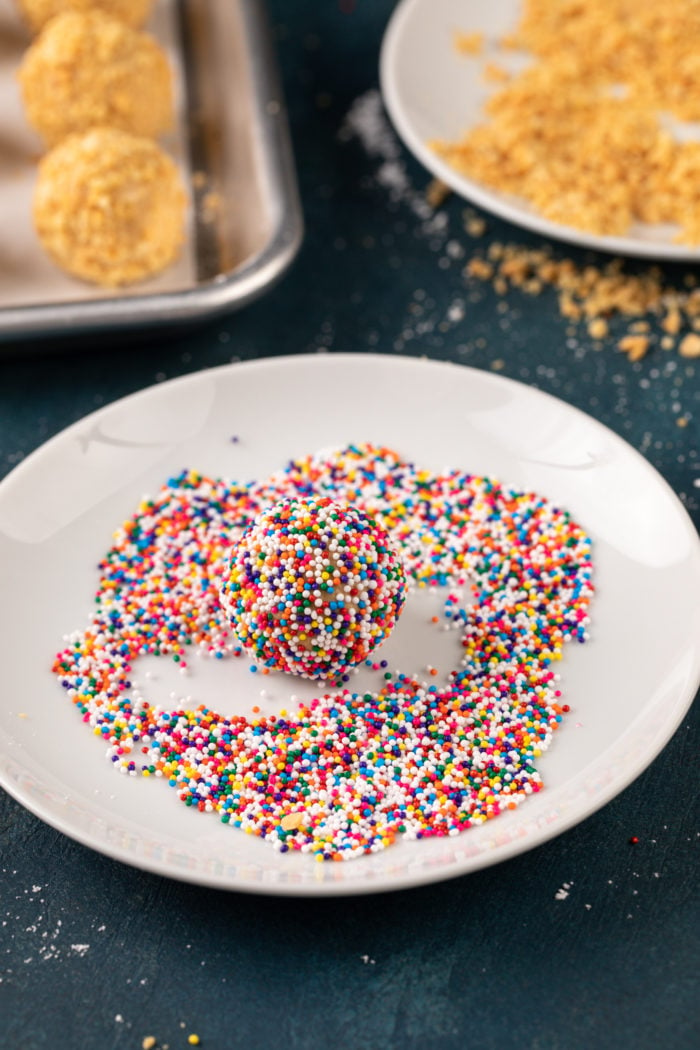 Peanut butter ball being rolled in rainbow nonpareil sprinkles on a white plate.