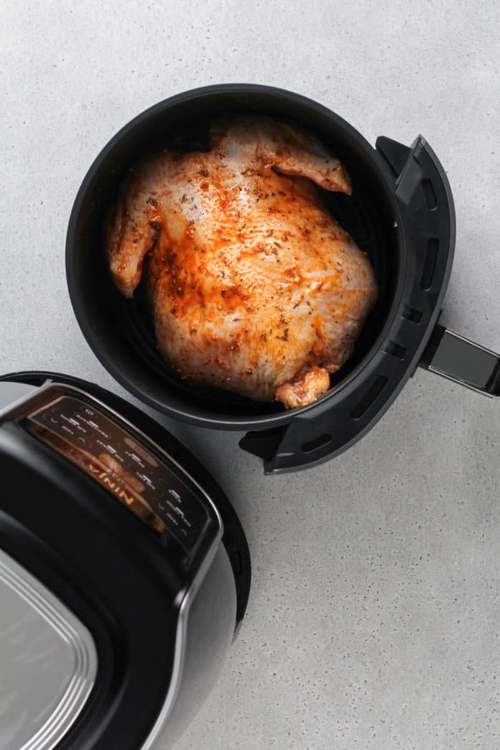 Whole chicken rubbed with seasoning placed breast-side down in the basket of an air fryer.