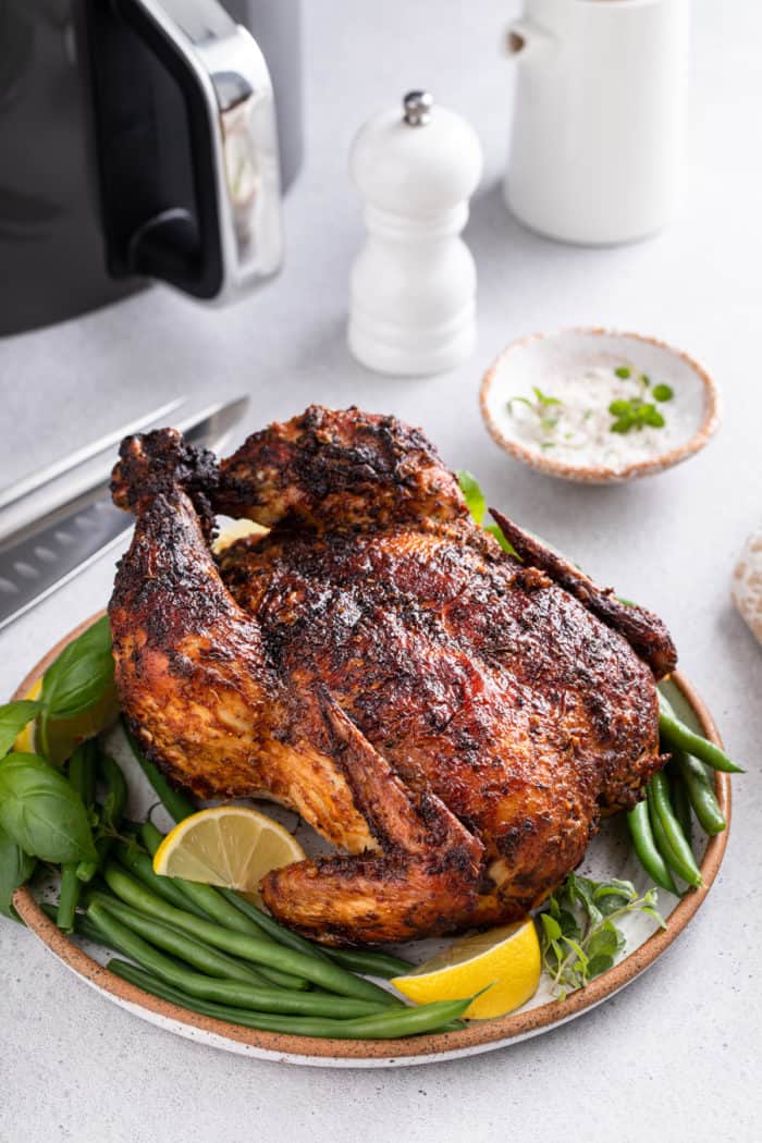 Whole air fryer rotisserie chicken set on a platter with herbs, green beans, and lemon wedges. An air fryer is visible in the background. 