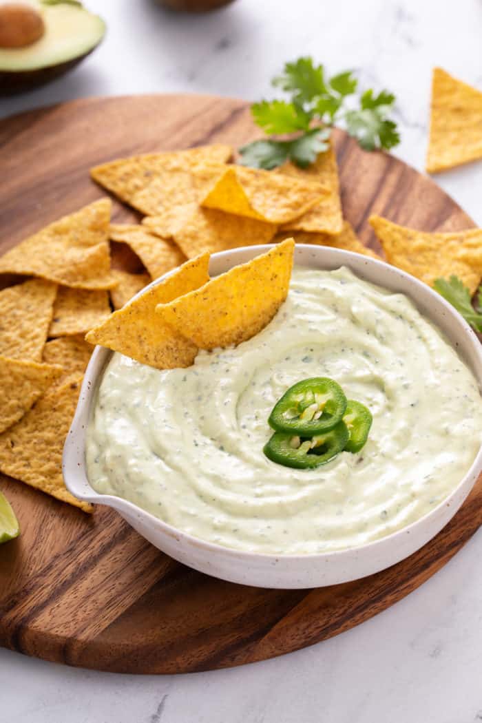 White bowl filled with jalapeño dip, with two chips in the dip. The bowl is on a wooden platter surrounded by tortilla chips.