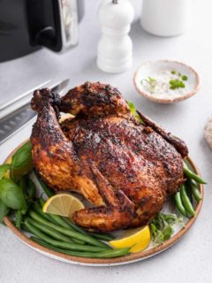 Whole air fryer rotisserie chicken set on a platter with herbs, green beans, and lemon wedges. An air fryer is visible in the background.
